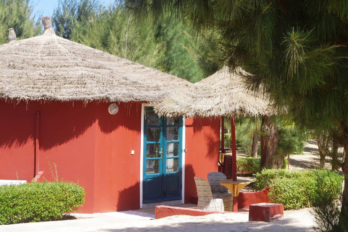 A red building with a blue door, thatched roofs, and a small patio area with outdoor seating.