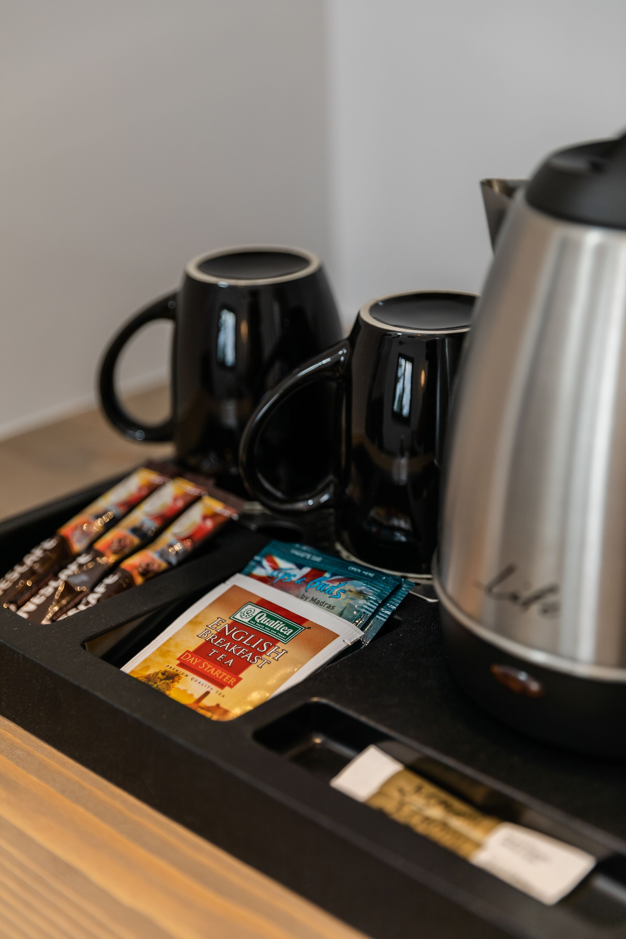 Un plateau de service à café comprenant deux tasses noires, une cafetière en argent et plusieurs sachets de thé.