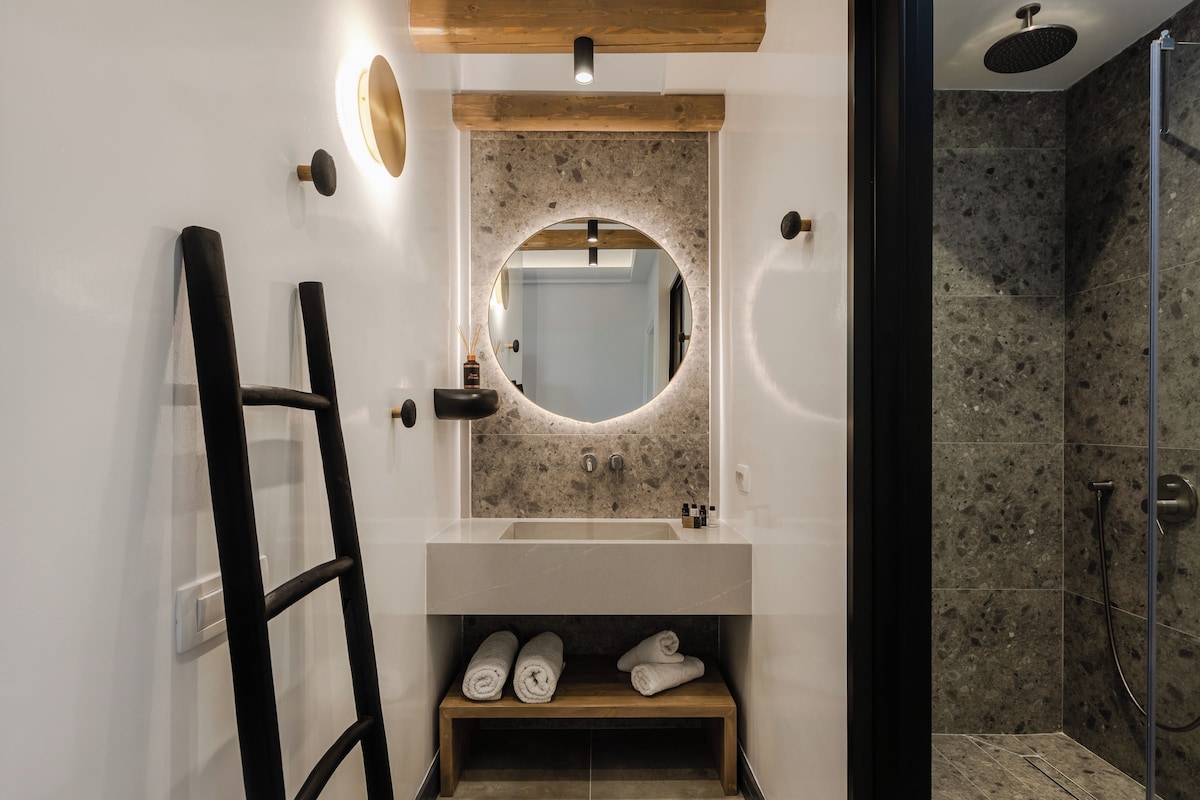 A modern bathroom with a round mirror, a wooden shelf with towels, and a shower area.