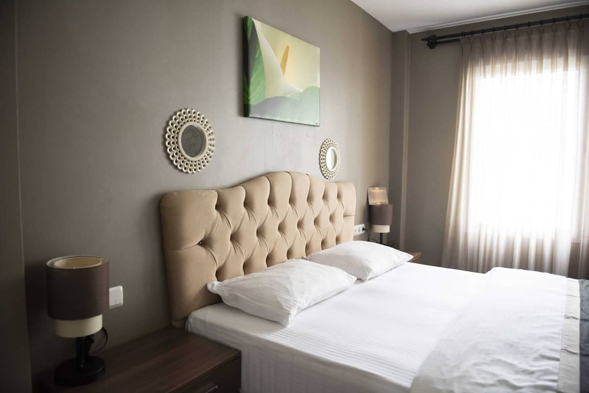 Een net opgemaakt bed met witte lakens, een gestoffeerd hoofdeinde en twee bedlampjes.