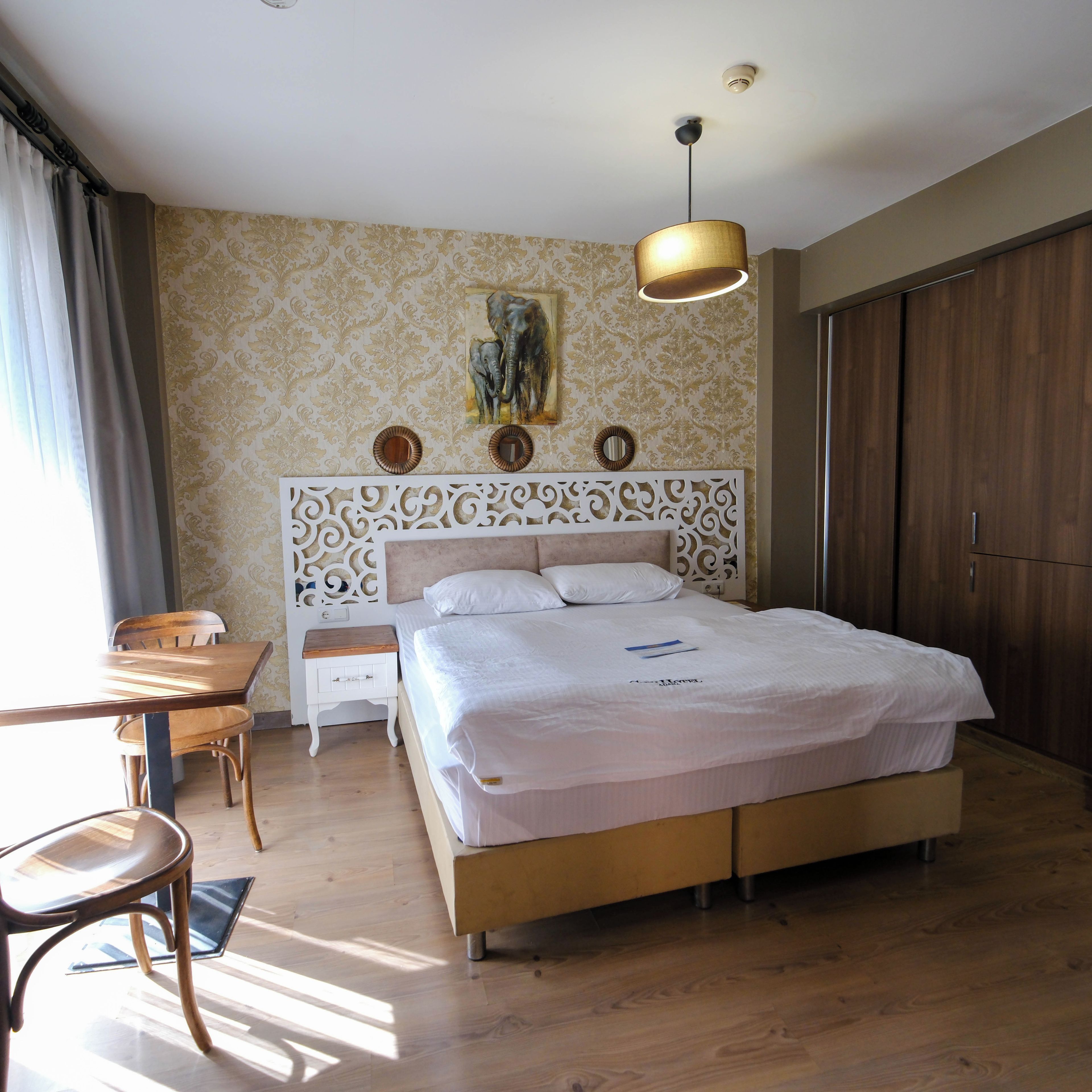 A hotel room with a large bed, a desk, and a chair. There is a painting on the wall and a wardrobe.
