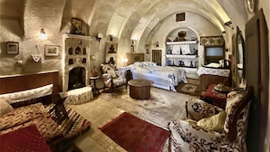 Una habitación con un techo alto abovedado, una chimenea, una cama con cubrecama blanco y una mesa con lámpara.
