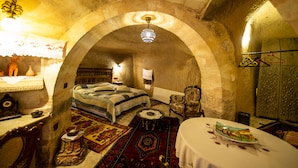 Una habitación con un arco de piedra, una cama con cubrecama estampado, una silla, una mesita con un libro y una lámpara colgante.