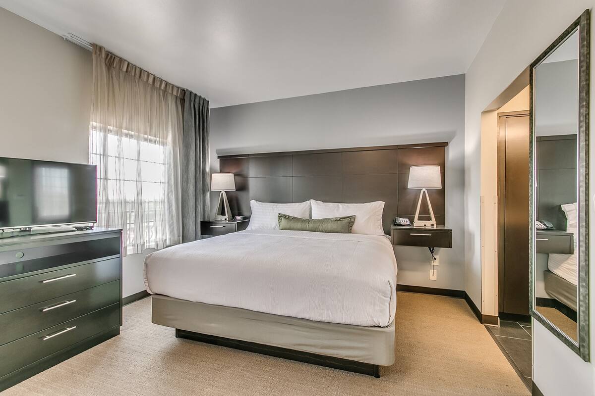 A modern hotel room with a large bed, a flat-screen TV, two bedside lamps, a dresser, and a large mirror.