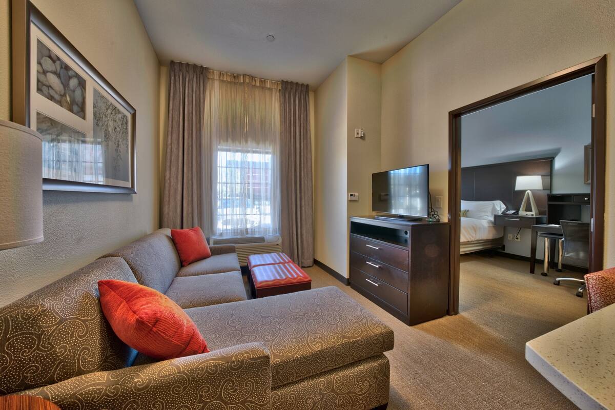 A hotel room with a sofa, ottoman, a TV, a desk, and a bed.