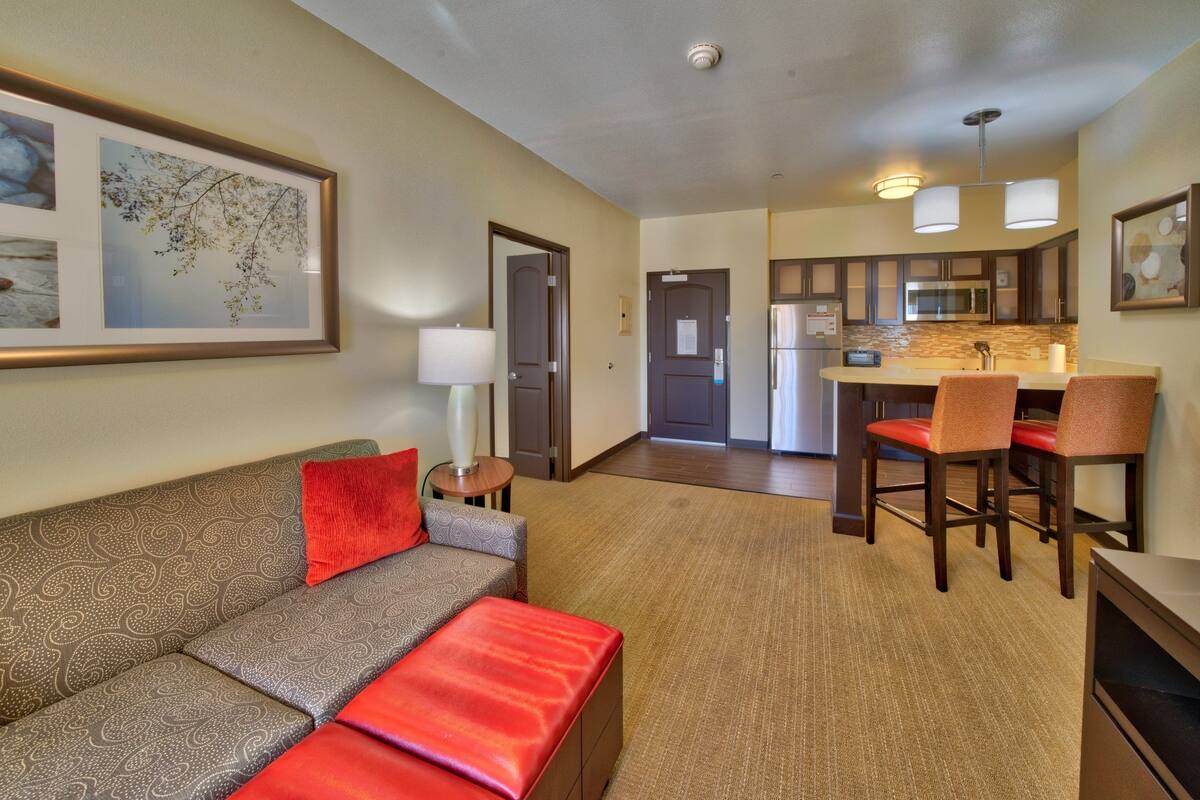 A modern hotel room with a sofa, a red throw, a dining area with chairs, a kitchen with a microwave, and framed artwork on the walls.