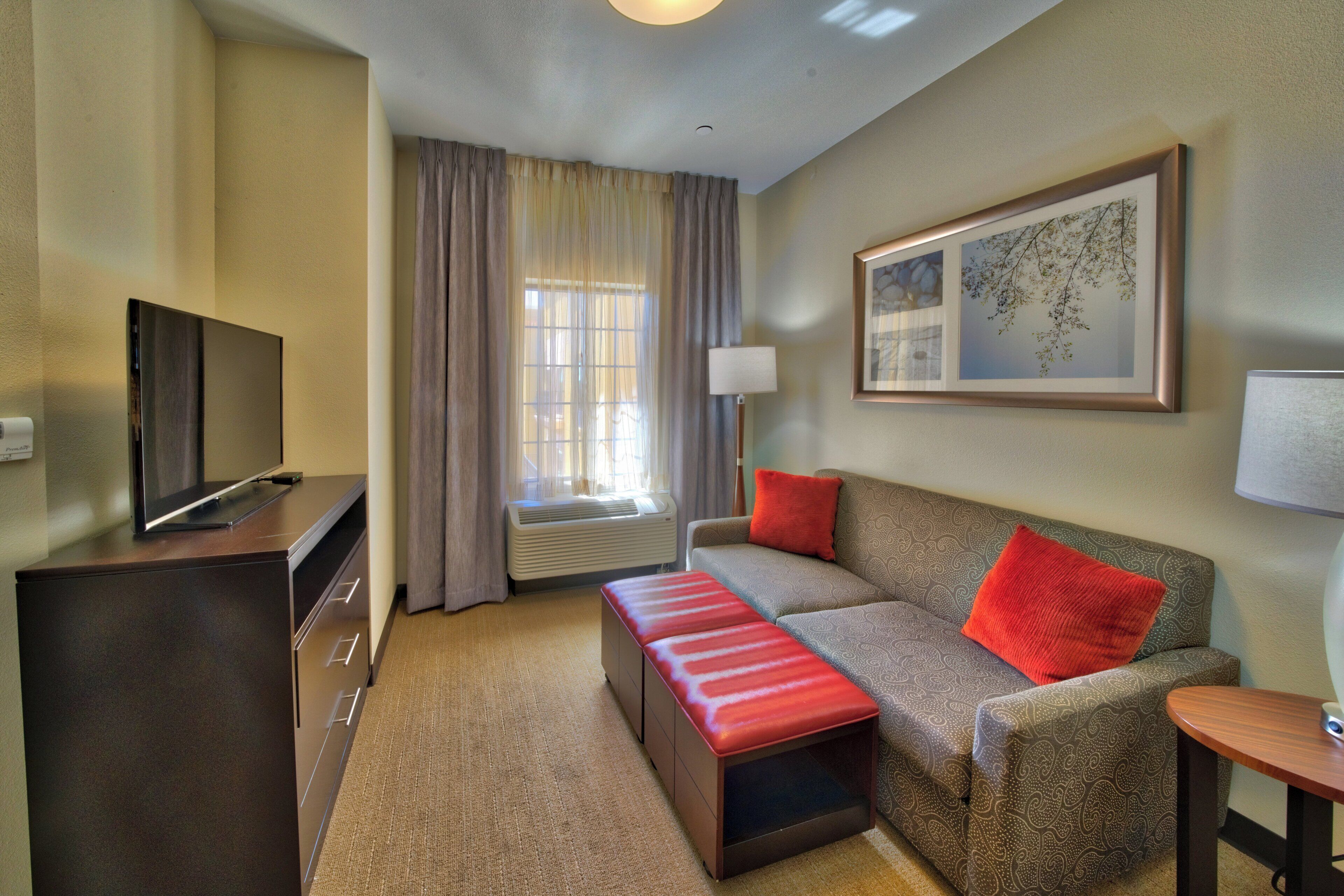 A hotel room with a flat-screen TV, a sofa with red cushions, a small table, a lamp, and a window with curtains.