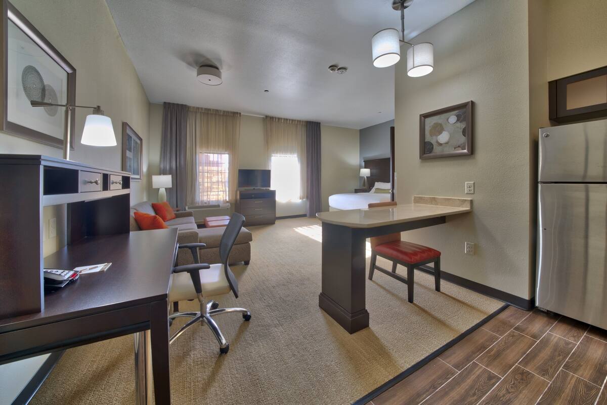 A modern hotel room with a kitchenette, a dining area, and a seating area with a sofa and chairs.
