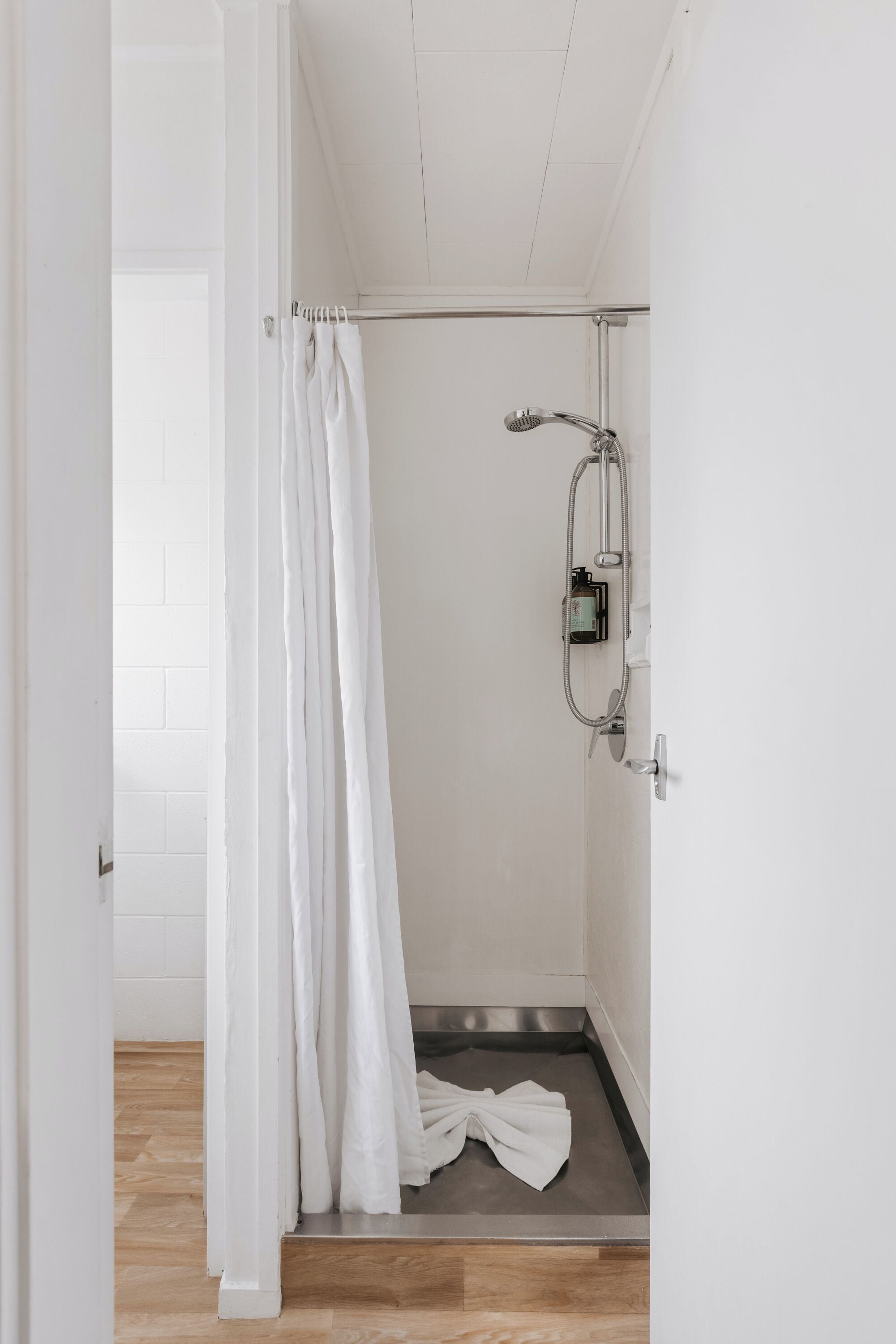 A shower area with a white curtain, a handheld showerhead, and a bottle of shampoo.