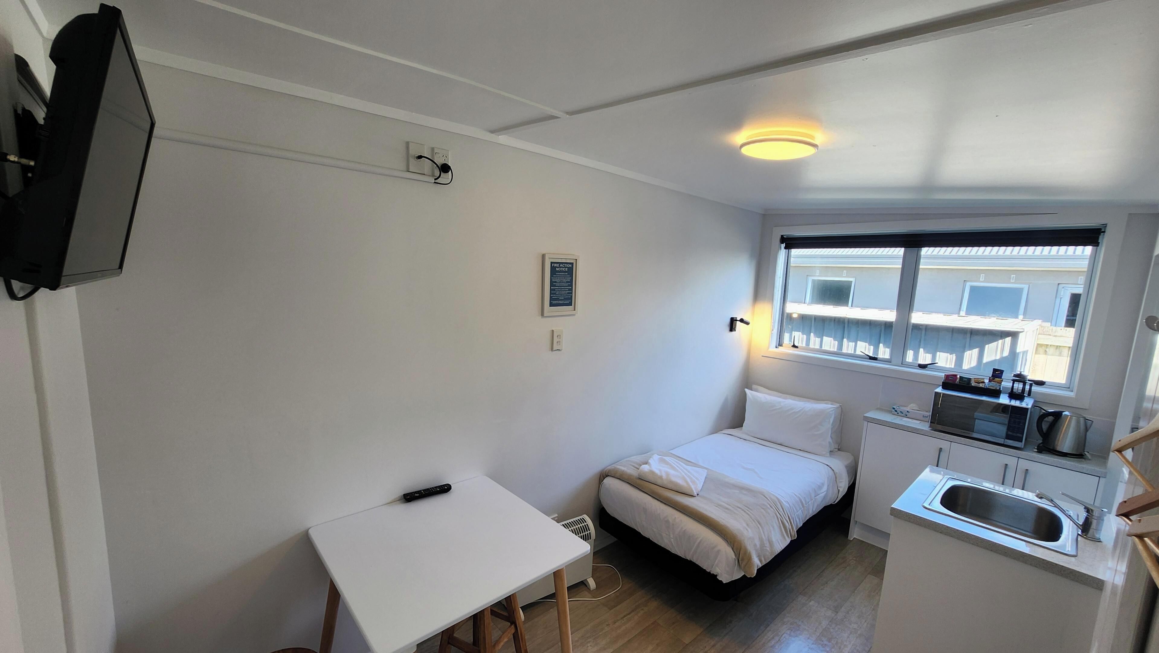 Una habitación compacta con cama, una mesita, kitchenette y ventana con vistas a edificios.