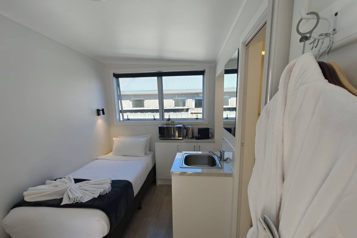 A compact hotel room with a bed, a small kitchenette, a sink, and a window with a view of buildings.