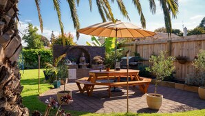 A patio with a wooden table and benches, a barbecue grill, and a large umbrella. - Roselands Motel (Tauranga)