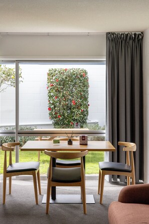 Un comedor moderno con una mesa y sillas de madera, una planta en maceta con flores rojas, y vistas a un edificio blanco a través de amplias ventanas.