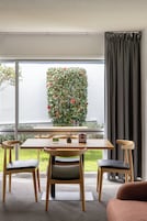 Un comedor moderno con una mesa y sillas de madera, una planta en maceta con flores rojas, y vistas a un edificio blanco a través de amplias ventanas.