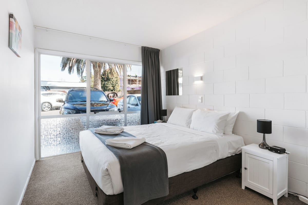 A hotel room with a bed, a nightstand, a window with a view of parked cars, and a white brick wall.