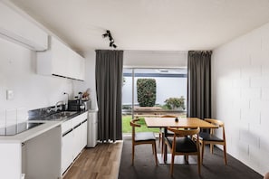 A compact kitchen with a dining area, a view of a garden, and a white brick wall. - Roselands Motel (Tauranga)