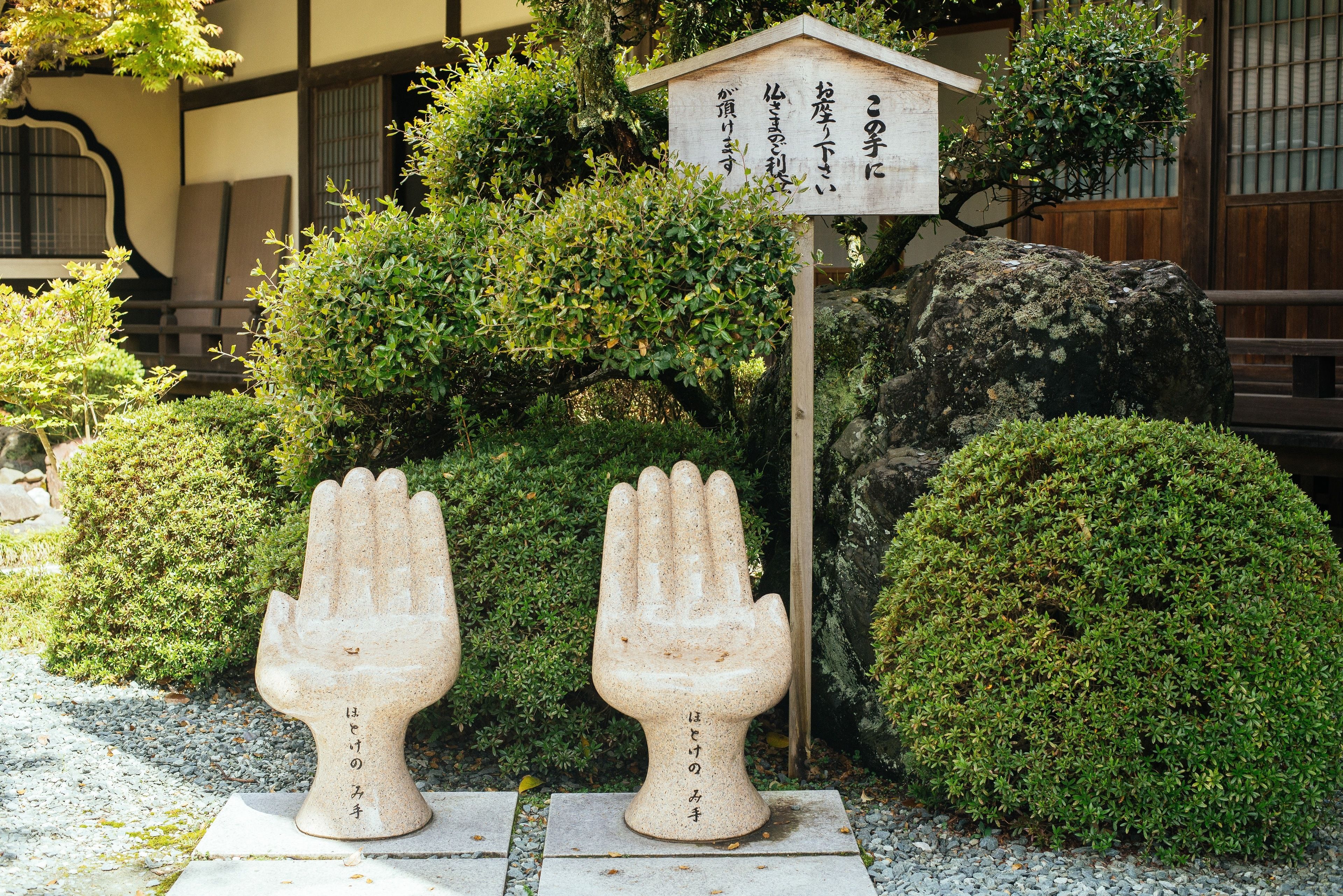 Kivestä veistetty käsiä esittävä patsas, jossa on japaninkielistä tekstiä. Patsasta ympäröivät hyvin hoidetut pensaat, ja taustalla näkyy perinteinen rakennus.