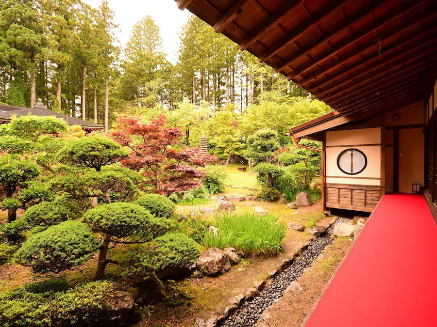 Un jardin japonais traditionnel avec un chemin rouge, des lanternes en pierre et un bâtiment en bois doté d’une fenêtre circulaire.