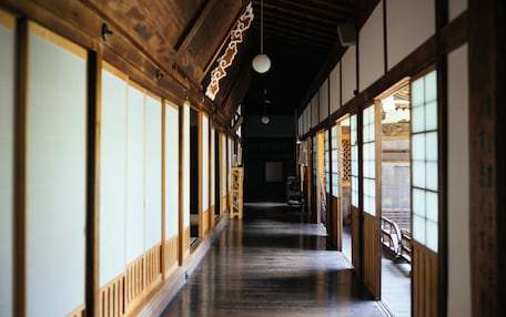 A traditional Japanese corridor with wooden beams, tatami flooring, sliding doors, and a hanging light fixture.