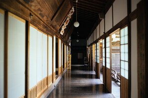Un pasillo japonés tradicional con vigas de madera, suelo de tatami, puertas correderas y un aplique colgante.