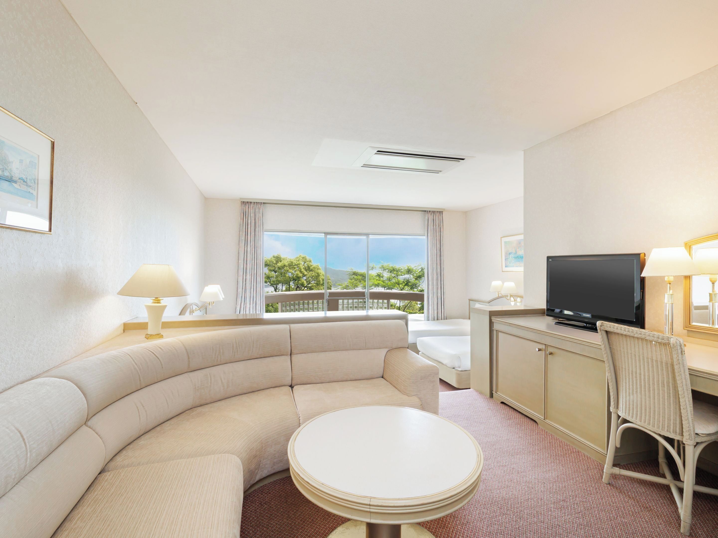 A hotel room with a curved sofa, a round table, a TV, and a balcony with a view of trees and mountains.