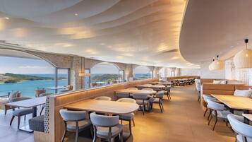 A modern restaurant with a view of the sea, featuring a bar area, seating arrangements, and contemporary lighting fixtures.