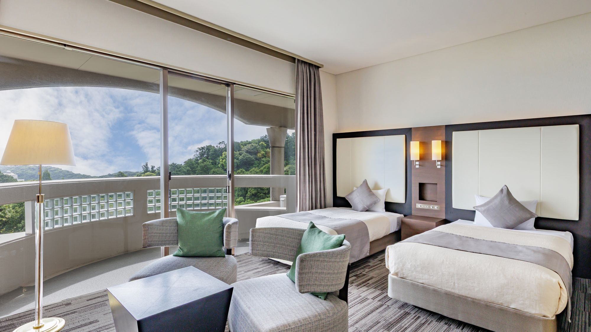 Habitación de hotel con dos camas, un sofá, una mesita y un ventanal amplio que da a vistas de follaje.