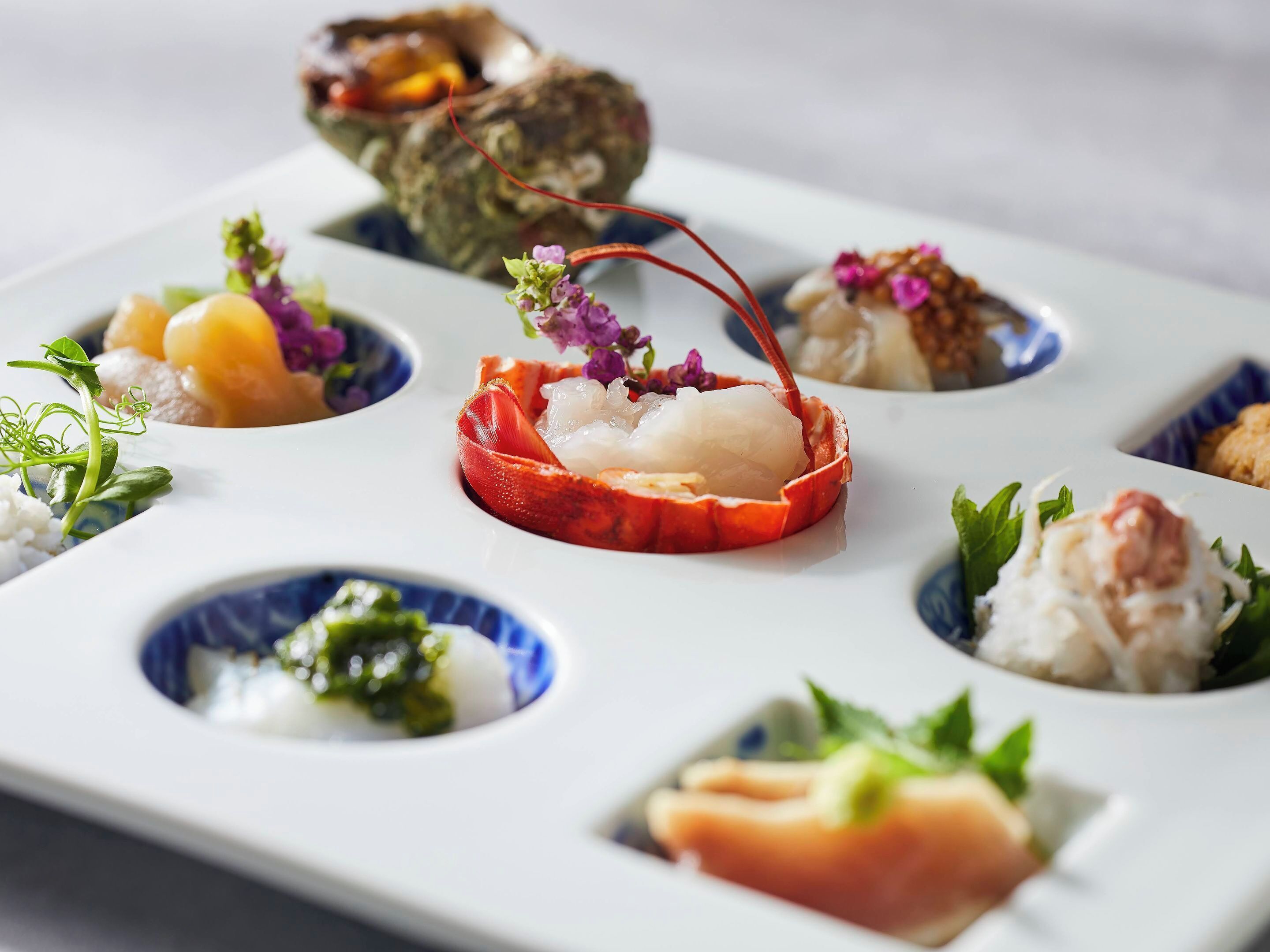 A white rectangular plate with multiple small dishes containing various food items, each garnished with small flowers and greens.