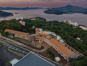 An aerial view of a coastal building complex with multiple structures, including a prominent tower, surrounded by lush greenery and a body of water.