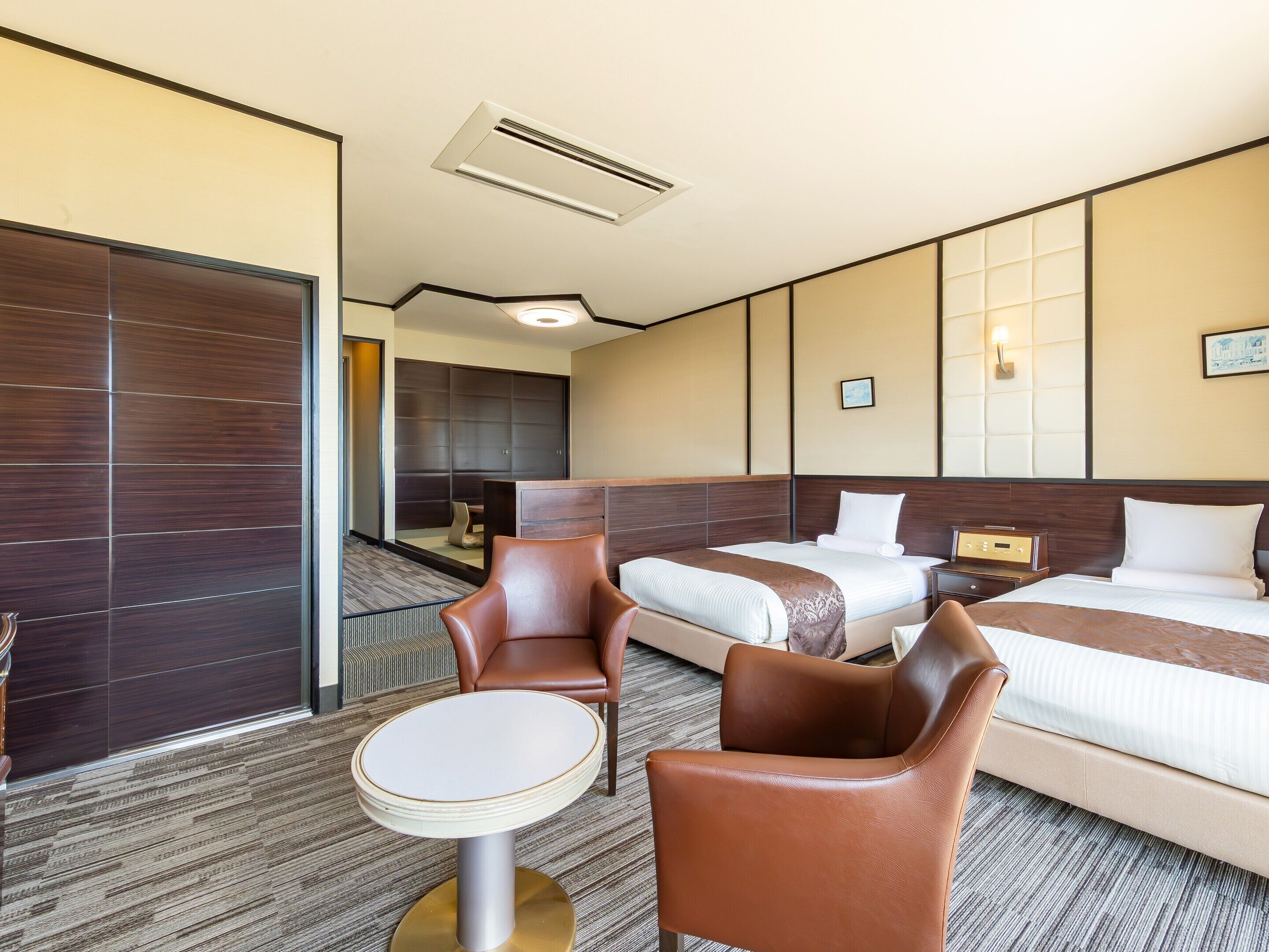 A hotel room with a bed, two brown leather chairs, a small round table, and a nightstand with a lamp.