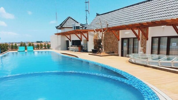 A swimming pool with a tiled edge, a covered patio area, and a building with a tiled roof. - G'Sign Style (Tuban)