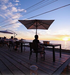 Une terrasse en bois, avec des tables et des chaises, offrant une vue sur la mer au coucher du soleil.
