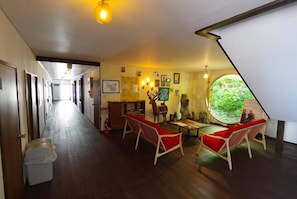 Un'hall d'albergo con un lungo corridoio, pavimento in legno, poltrone rosse imbottite, un'ampia finestra tonda che si affaccia sul verde, e diversi oggetti fissati alle pareti.
