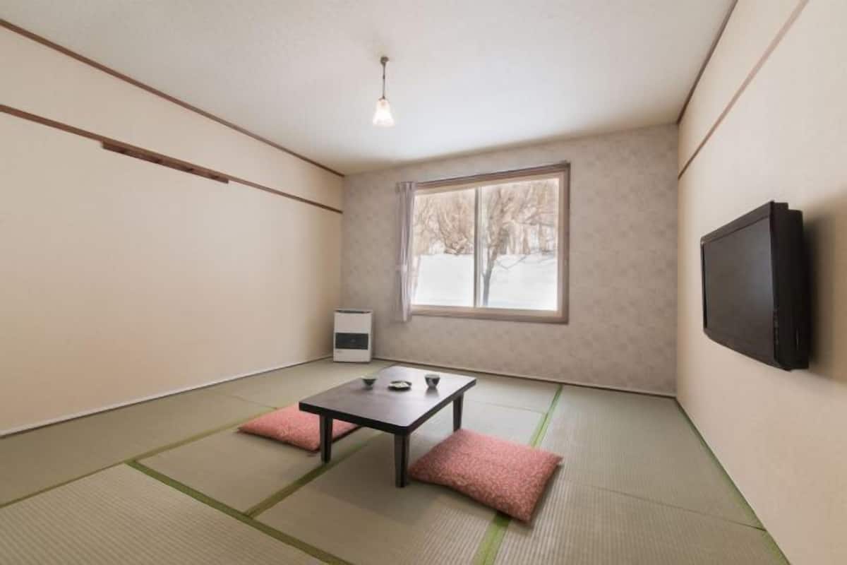 A room with tatami flooring, a low table with cushions, a heater, a window with a view of trees, and a mounted TV.