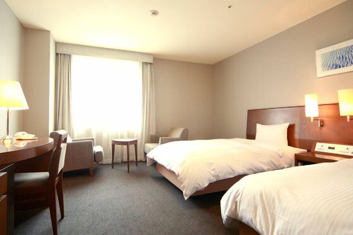Ein Hotelzimmer mit zwei Betten, einem Schreibtisch, einem Stuhl, einem kleinen Tisch und einem Fenster mit Vorhängen.