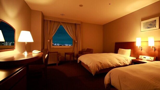 A hotel room with two beds, a desk, a chair, a window with curtains, and a city view. - Hotel Brillante Musashino (Saitama)