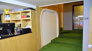 A room with a wooden desk, computer, and shelves with various items. There is a green carpeted staircase leading to another room.