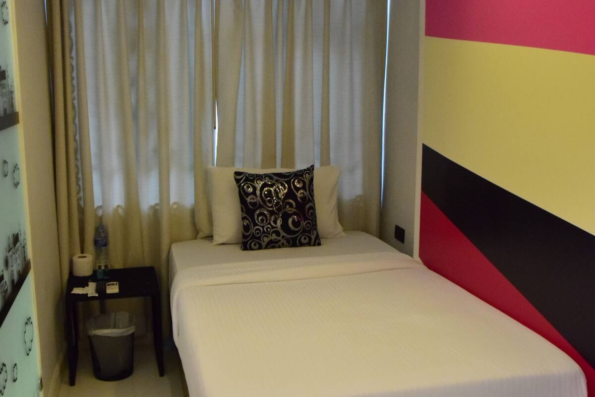A neatly made bed with a white comforter and two pillows, a bedside table with a lamp, a trash bin, and a wall with a bold color pattern.