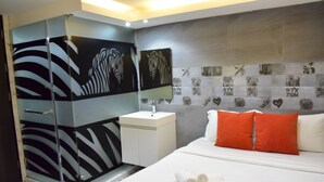 A modern hotel room with a glass shower enclosure, a white bedside table, a bed with white bedding and a red pillow, and a wall decorated with heart-shaped tiles.