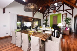A spacious dining area with a wooden table and chairs, a large woven light fixture, and a view of the outdoors through a sliding glass door. - Sankhara Beachfront Villas (Poste Lafayette)