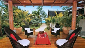 An outdoor seating area with wicker chairs and a red table, surrounded by tropical plants and a view of the ocean. - Sankhara Beachfront Villas (Poste Lafayette)