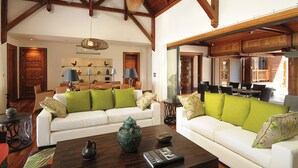 A spacious living room with a high ceiling and exposed wooden beams, featuring two white sofas with green cushions, a coffee table with decorative items, and a dining area with a wooden table and chairs. - Sankhara Beachfront Villas (Poste Lafayette)
