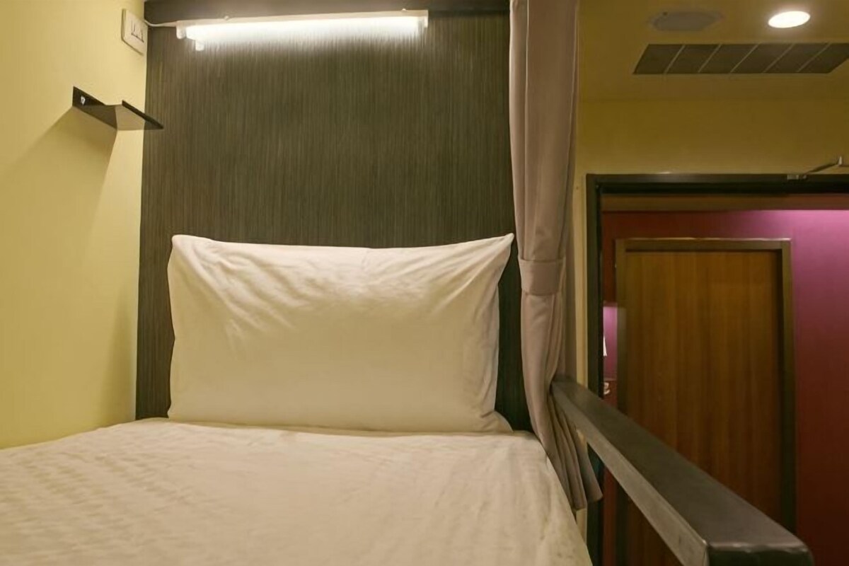 Un lit simple avec une tête de lit, une étagère fixée au mur et une porte visible au fond.
