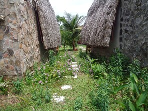 A stone-walled courtyard with thatched-roof structures, a stone pathway, and lush greenery.