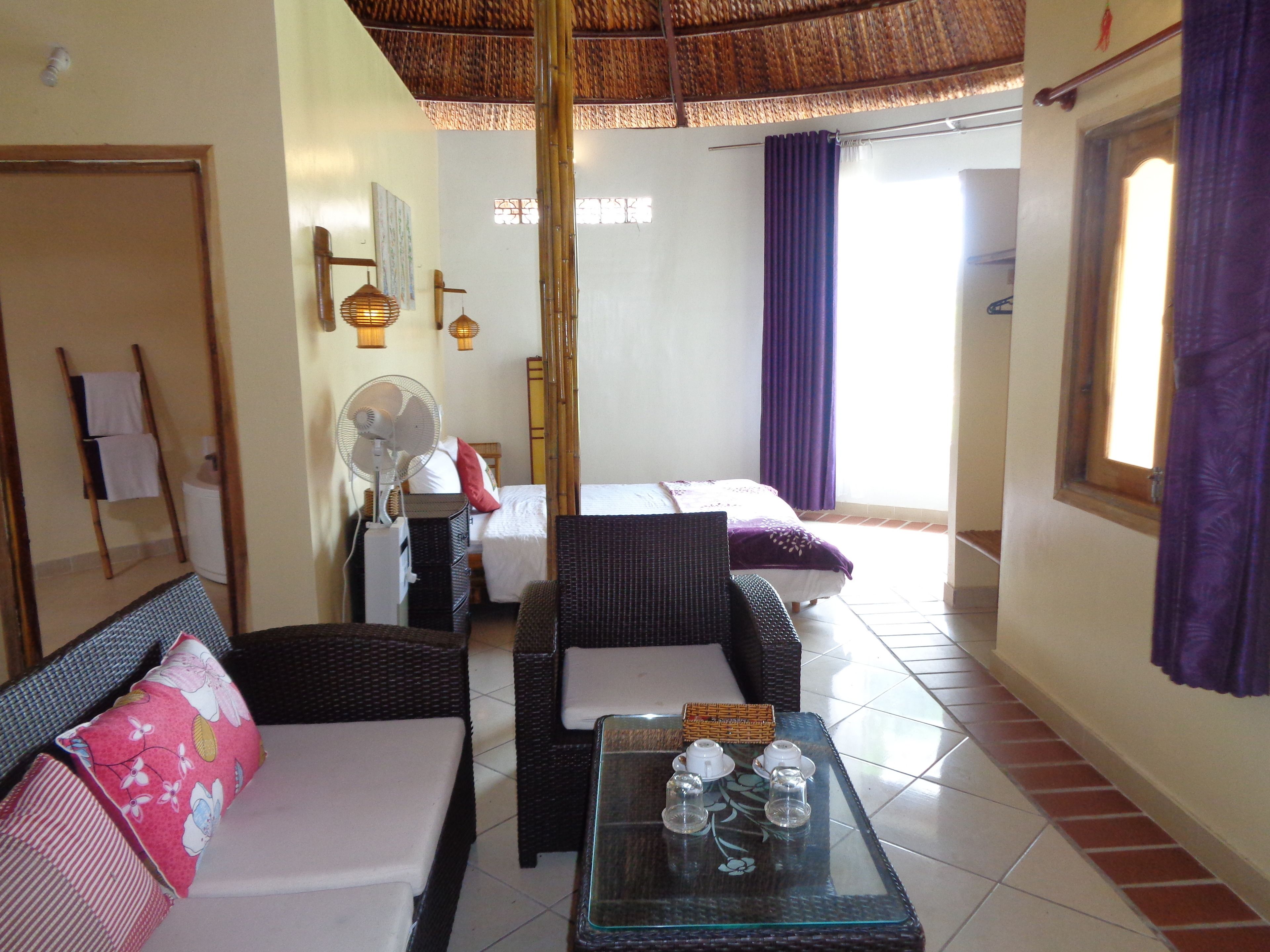 A hotel room with two beds, a sitting area with wicker furniture, a fan, and a table with glasses and a bottle.