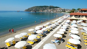 Une plage bordée de rangées de transats et de parasols, une mer d’un bleu limpide et, au loin, une colline.