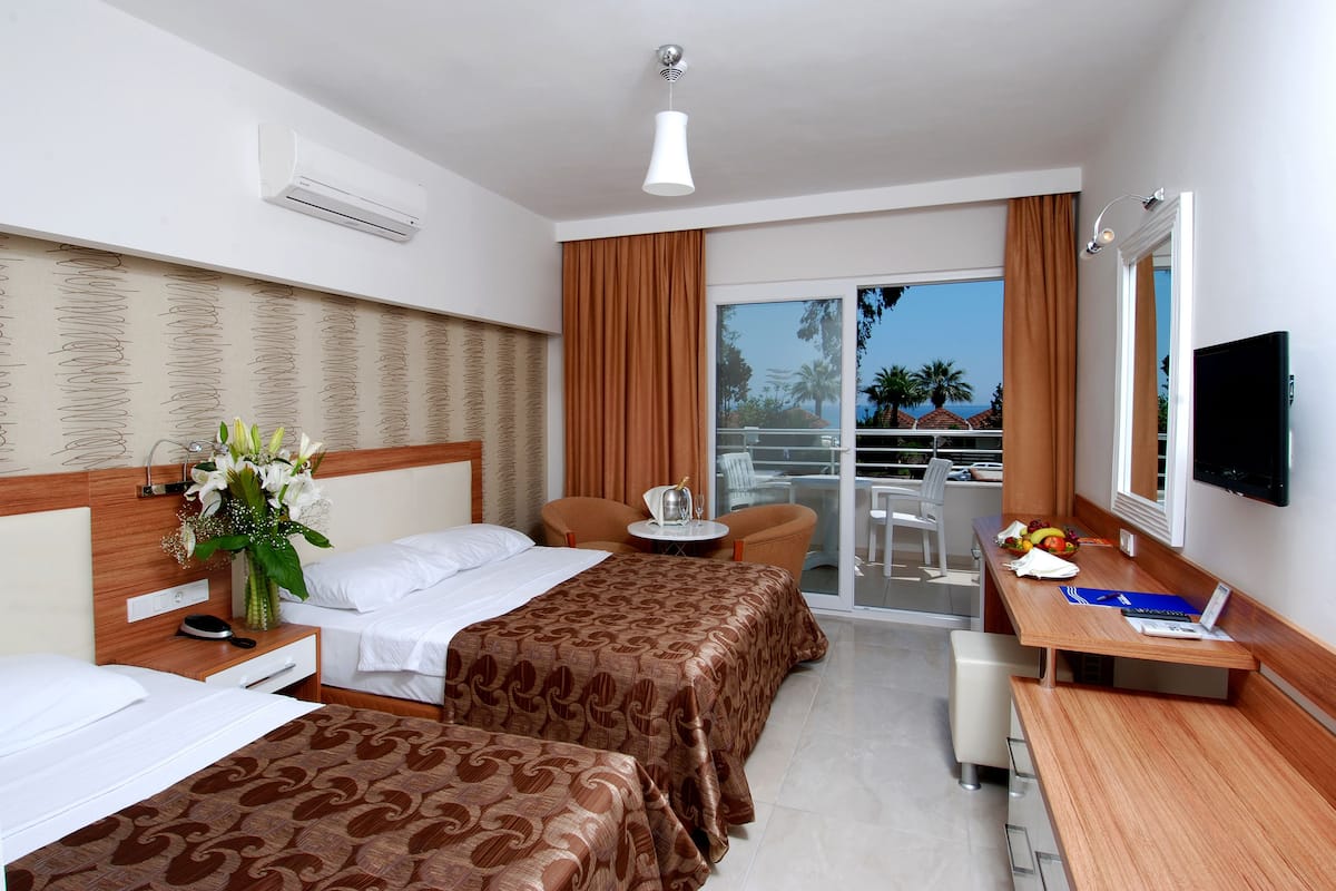 A hotel room with two beds, a desk, a chair, a TV, and a balcony with a view of palm trees.
