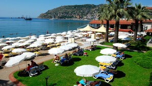 Un complexe hôtelier situé en bord de mer, doté de nombreuses chaises longues et de parasols, d’un espace piscine et offrant une vue sur la mer et une colline en arrière-plan.
