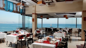 Un restaurant avec des tables dressées pour le service, de grandes fenêtres donnant sur la mer et des ventilateurs de plafond.