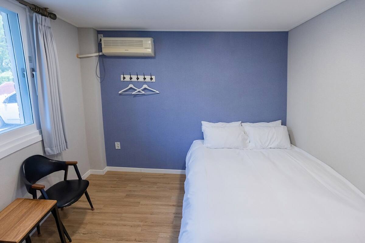 Een slaapkamer met een bed, een stoel, een tafel, een airconditioning en een kapstok.